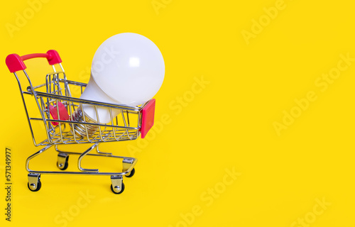 Shopping trolley and an energy-saving light bulb on a yellow background. Favorable purchase of electric power equipment to save costs. Saving energy and caring for the environment. Free space for text
