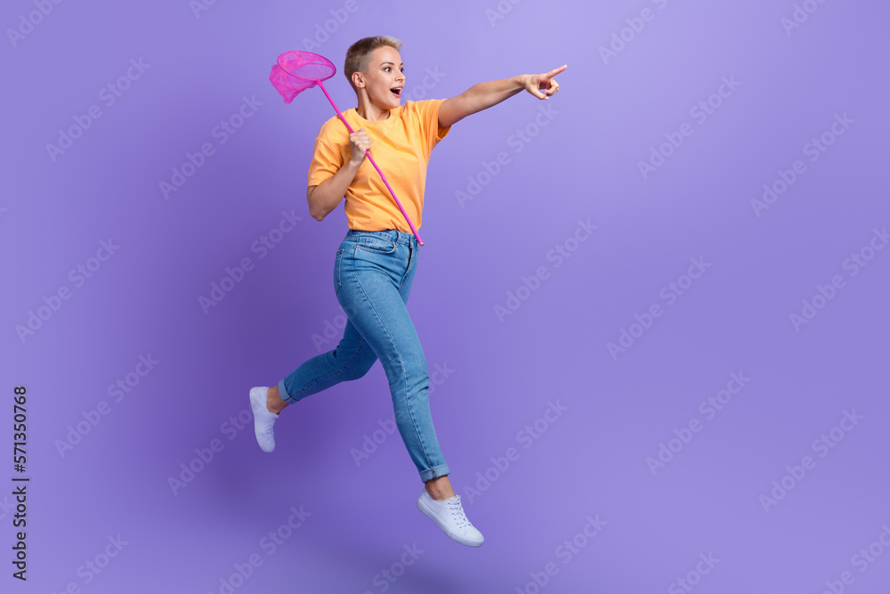 Full size photo of optimistic excited girl sneakers denim pants hold net directing empty space isolated on purple color background