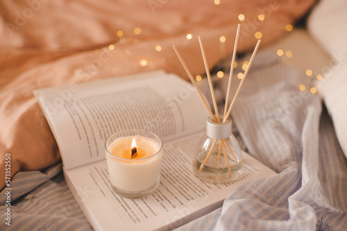 Fotografia Burning scented candle with glass bottle with home liquid perfume on paper book in bed closeup over glowing lights in bedroom