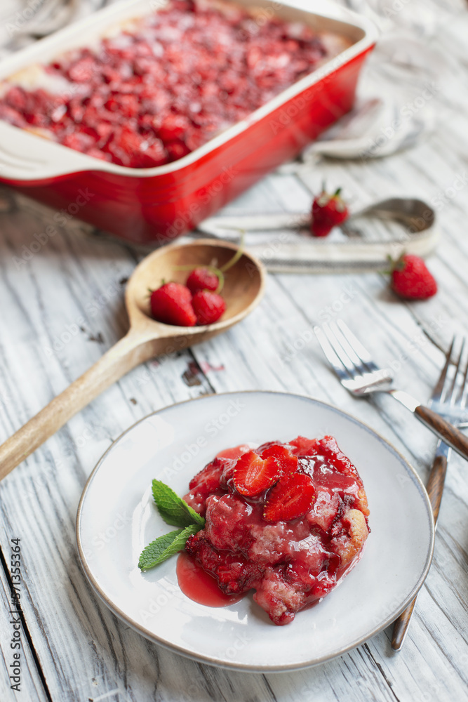 High angle view of sweet homemade strawberry cobbler or Sonker baked in a red ceramic pan over a rustic white wood table. Extreme selective focus with blurred background. High angle view of sweet homemade strawberry cobbler or Sonker baked in a red ceramic pan over a rustic white wood table. Extreme selective focus with blurred background.