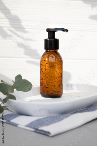 Amber bottle with facial cosmetics on a tray with a branch of eucalyptus on the gray wooden background, facial cleaner. Front view