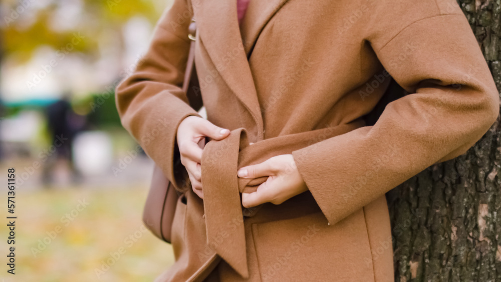 Close-up hands of girl tie a belt on brown coat in an autumn park