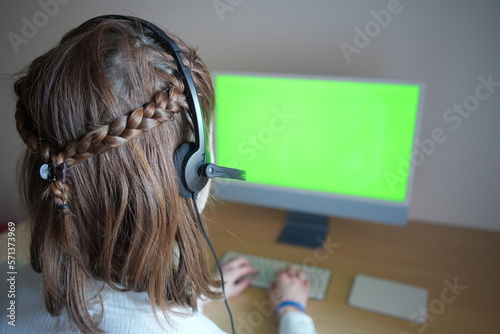 Woman wearing headset is sitting at a desk and typing on a computer with a green screen. Her face is focused on the monitor as she works on a project, and her hands are positioned on the keyboard. 