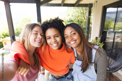 Portrait of happy diverse teenage female friends embracing on balcony taking selfie