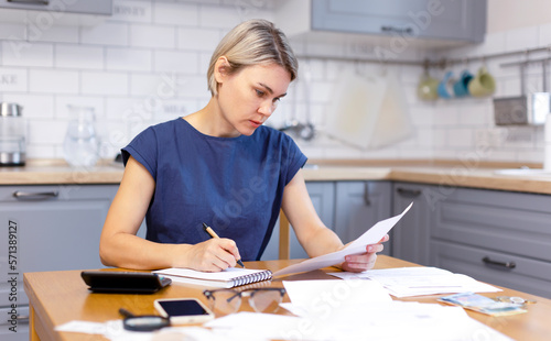 A woman works at home with business papers, counts money and writes down information. The accountant works with invoices and taxes. A blonde woman in a blue dress is in business.