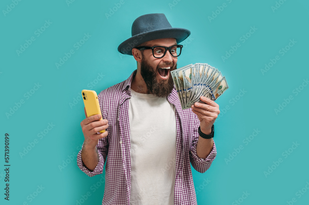 Excited happy young male winner feeling joy while holding money won, winning lottery, placing bets, getting cashback online gift on blue background. Human face emotions and betting concept. - obrazy, fototapety, plakaty 
