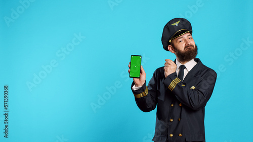 Positive aircrew captain showing phone with greenscreen, holding smartphone with isolated display and chroma key template. Young airplane pilot in uniform looking at mockup copyspace screen. photo