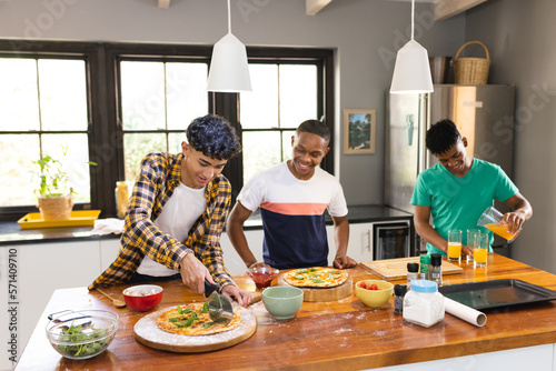 Happy diverse teenage male friends cutting self made pizza in kitchen