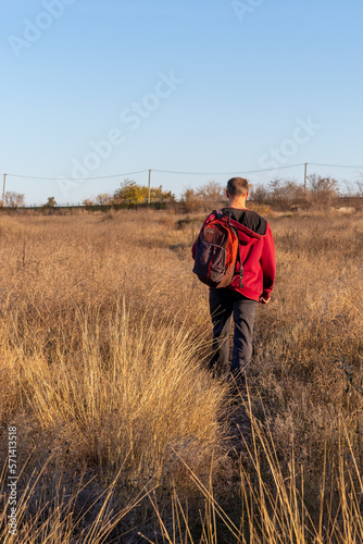 Man with backpack walks along path into distance, into field, in direction of railway. Back view. Concept of freedom and travel