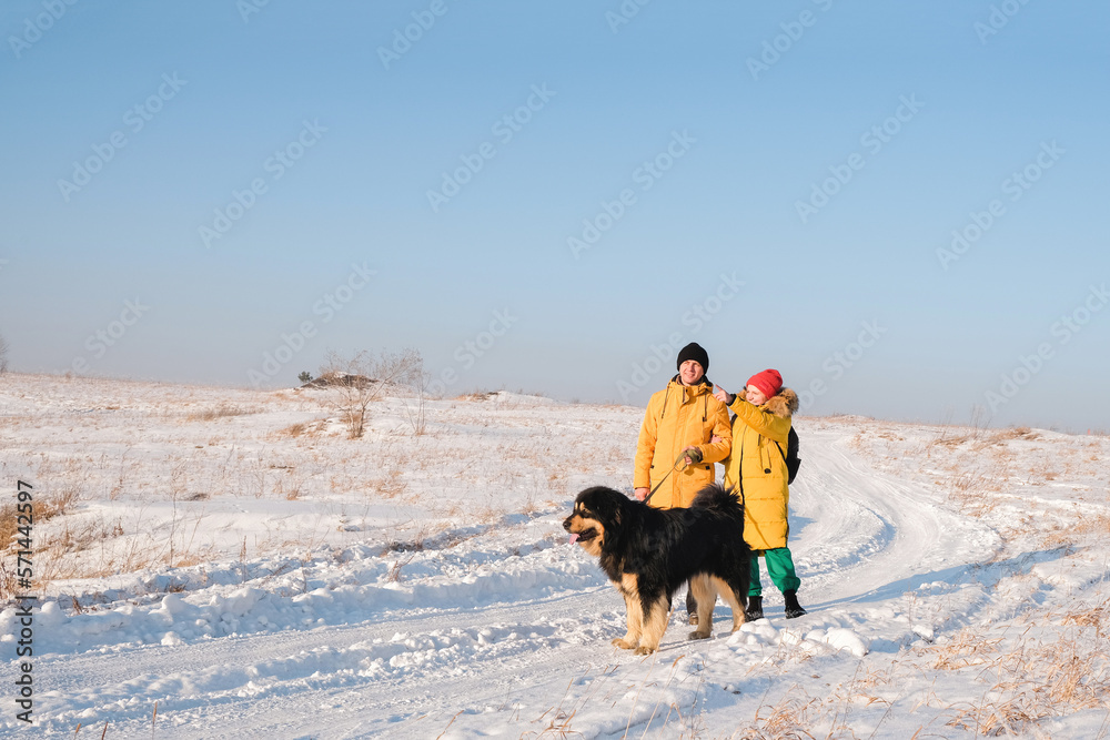 Male with female in yellow coats walking with  big black dog on winter background. Family winter activity with pet on sunny day outdoor. 