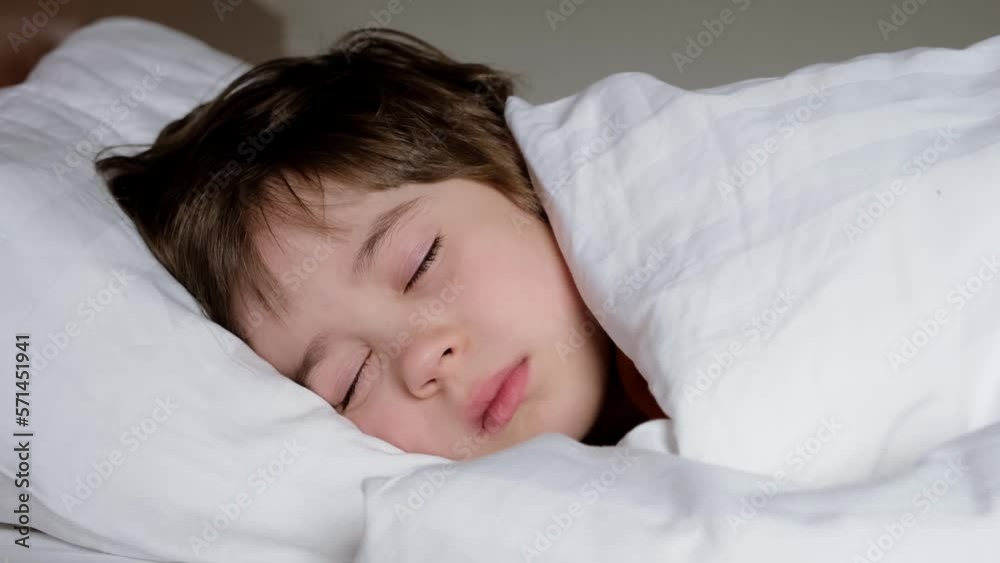 Smiling boy sleeps in bed under blanket. The child at daytime relax. White linens. Dreaming time. Concept of soft and comfortable mattress, soft pillow and warm blanket. The happy dream. Good morning. Smiling boy sleeps in bed under blanket. The child at daytime relax. White linens. Dreaming time. Concept of soft and comfortable mattress, soft pillow and warm blanket. The happy dream. Good morning.