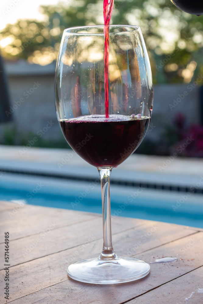 Crystal glass next to a swimming pool while it is being filled with red wine.