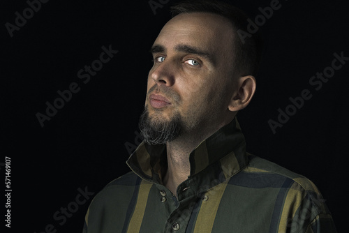 Beautiful bearded man in a studio in an unobtrusive way. Portrait of a sexy man in a checkered shirt against a black background. Dramatic lighting in soft light. Slightly side view.
