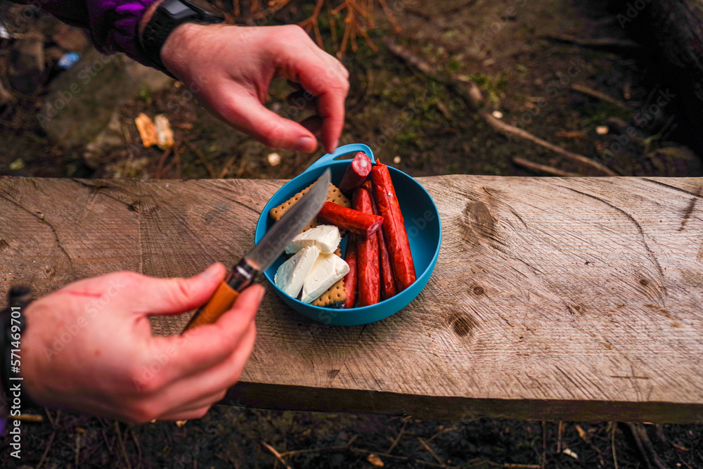 Obraz premium Tourist breakfast. Tourist food in a bowl. Knife in hand. Cooking lunch in nature. Snack while hiking. Person cuts the sausage.