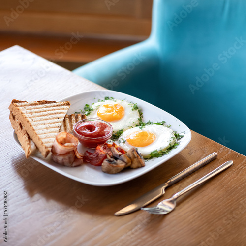 Breakfast at restaurant. Fried eggs with bakon  mushrooms  toast and sauce. Breakfast and cutlery on wooden table. Soft focus. Copy space. Side view.