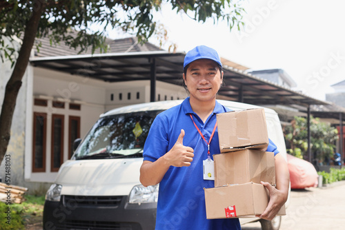 Young Delivery Man in Blue Uniform Carrying Pile of Cardboard Box and Showing Thumb-up In Front Of Truck