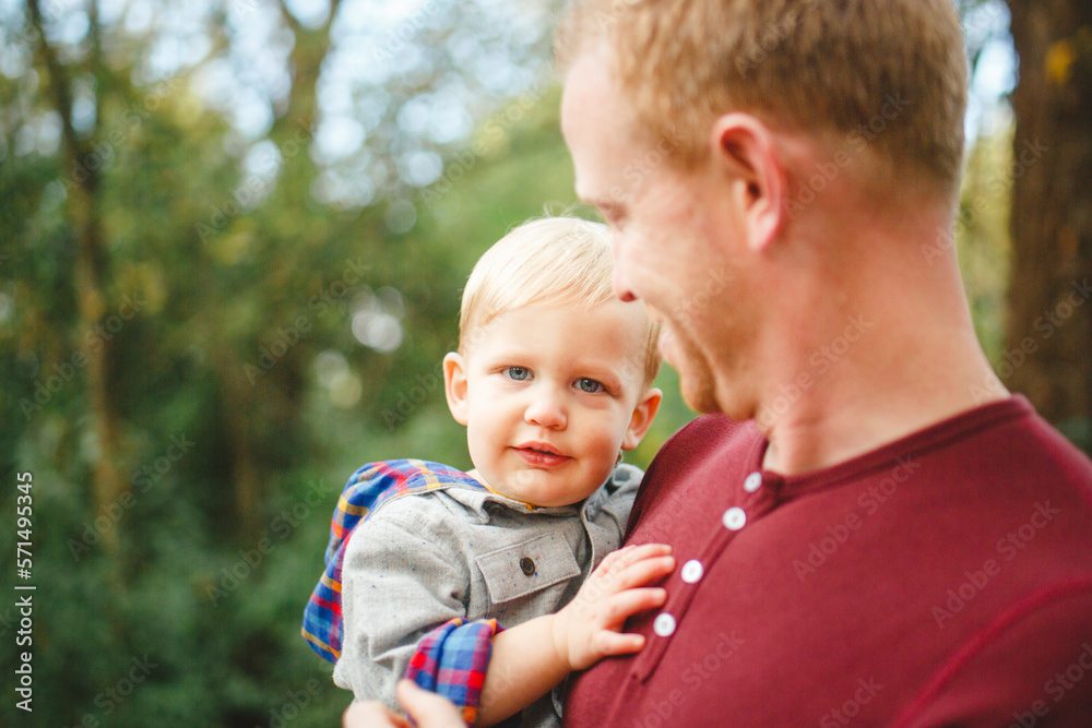 A small boy with direct gaze is held in his father's arms in a forest A small boy with direct gaze is held in his father's arms in a forest