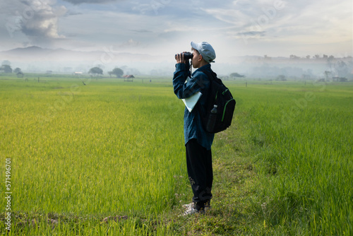 Asian boy in plaid shirt wears cap and has a backpack, holding a binoculars and map, standing on ridge rice field of asian farmers to observe fish and birds on tree branches and on sky, soft focus.