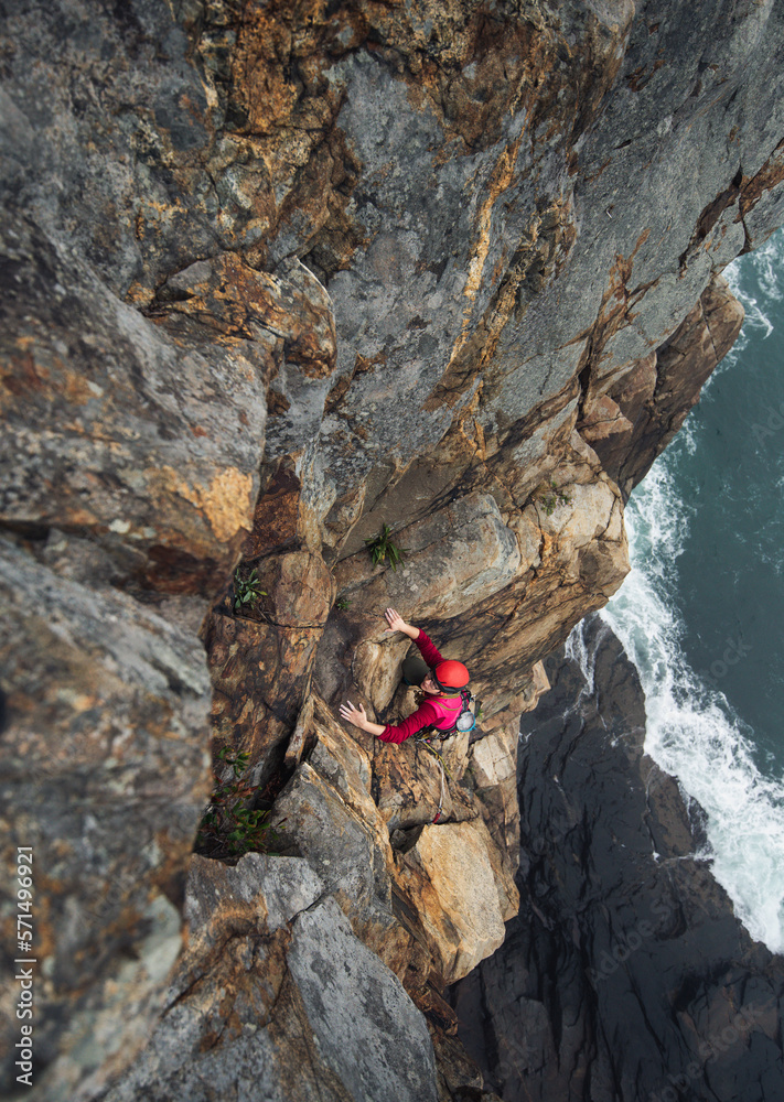 Woman wearing red trad climbing on lead with ocean below Woman wearing red trad climbing on lead with ocean below