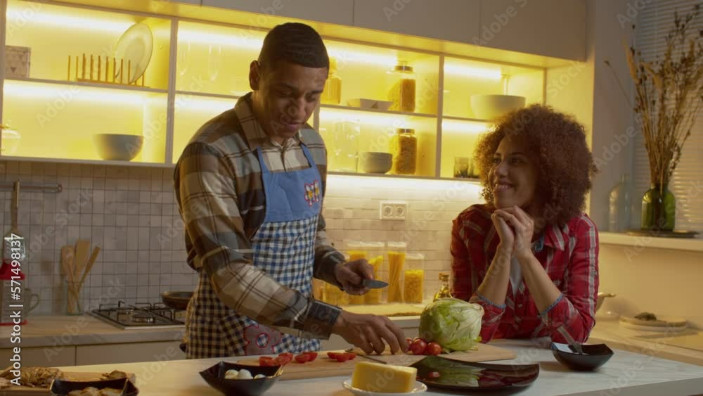 Handsome African American man in apron cooking fresh tasty salad, chatting with beloved pleased black woman while attractive couple enjoying leisure and preparing romantic dinner in kitchen.