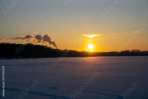 Sunset over the frozen surface of the river bay. The icy surface of the river covered with snow against the backdrop of the forest and the setting sun