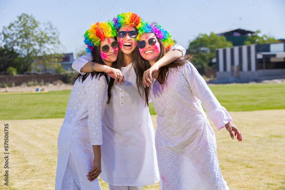 Group of happy young indian friends wearing white kurta and colorful hair wigs celebrating holi festival outdoor at park.