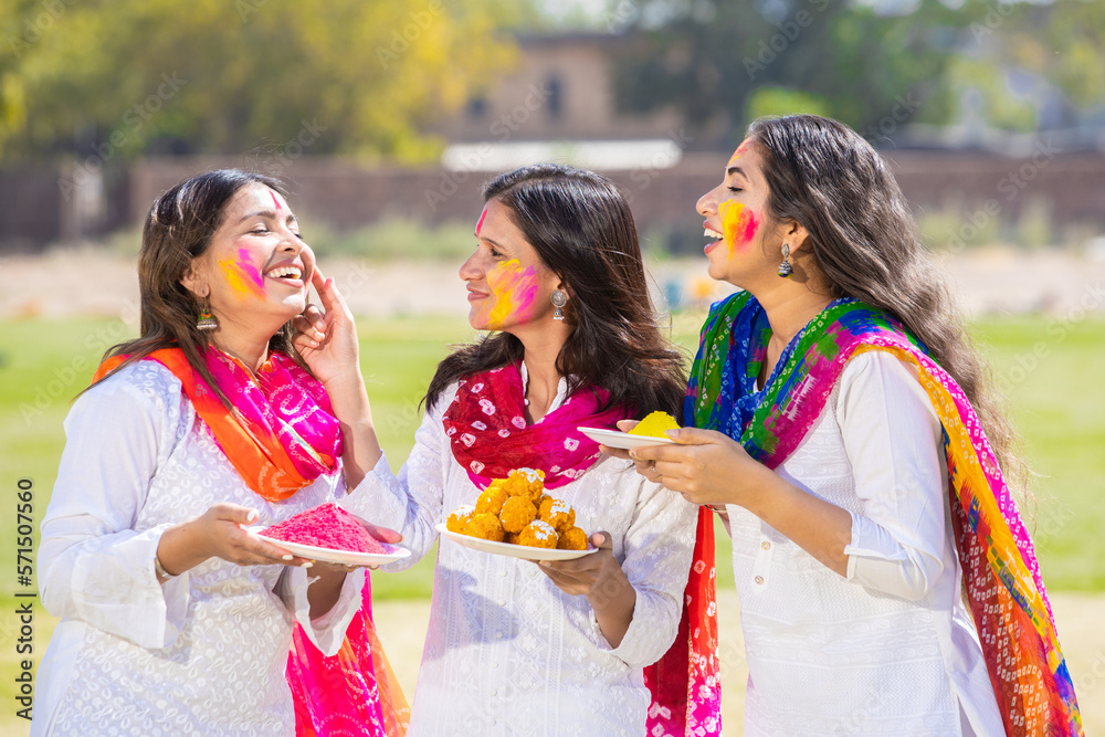 Fototapeta premium Group of happy young indian woman wearing white kurta dupatta outfit celebrating holi festival at park while holding plates full of gulal and laddoo sweet , woman having fun with colorful powder.