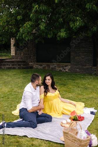 Beautiful couple sitting on a plaid on the grass. Elegant pregnant woman with husband waiting for baby. Future parents. Yeallow dress © Aleksandr