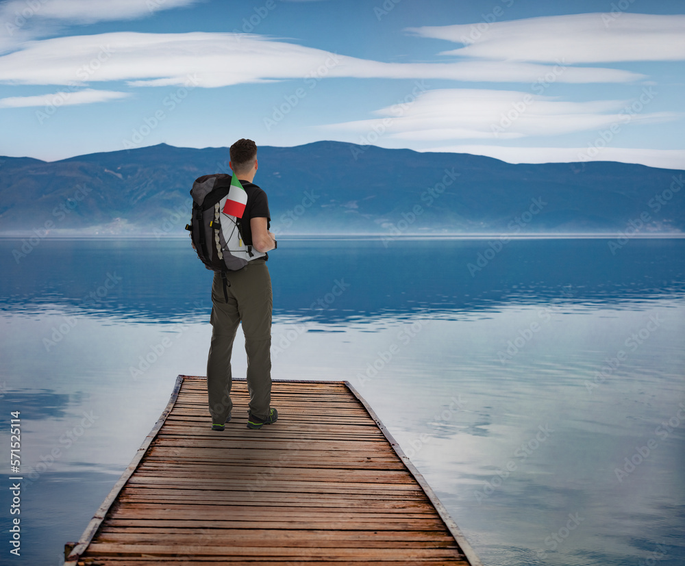 Еourist with a backpack and an Italian flag stands on a wooden pier on Lake Como. Splendid summer scene of Italy, Europe. Traveling concept background..