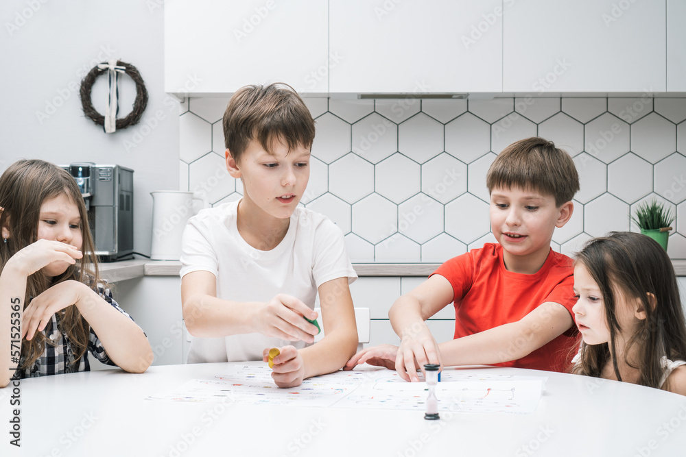 Happy little school friends play board game on paper map with dice and chips at kitchen table. Children throw dice and move figure. Home leisure, playtime. Entertaining interesting table game.