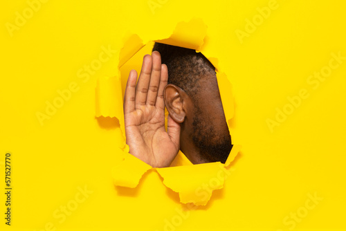 Eavesdropping. Unrecognizable black guy putting ear to torn yellow paper background, listening through hole, closeup