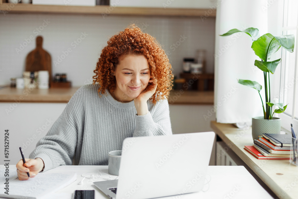 Cute girl with red curls watching online tutorial or lesson on laptop, smiling listening and looking attentively, making notes in copybook, sitting at kitchen table with smartphone and cup of coffee - obrazy, fototapety, plakaty 