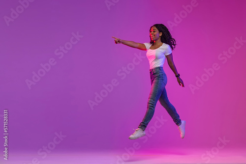 Look There. Happy Black Woman Jumping In Neon Light And Pointing Aside