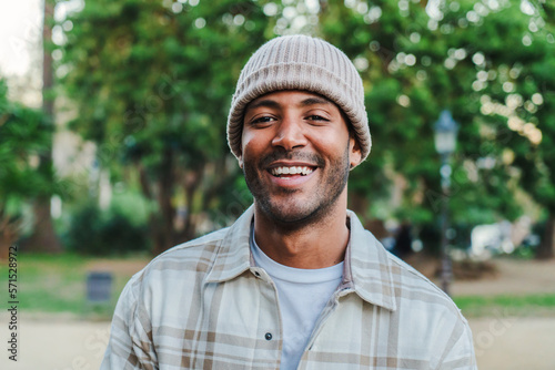 Close up portrait of young hispanic man with a beanie hat smiling and looking at camera outdoors. Front view of latin happy guy standing at park with carefree attitude. Lifestyle concept. High quality