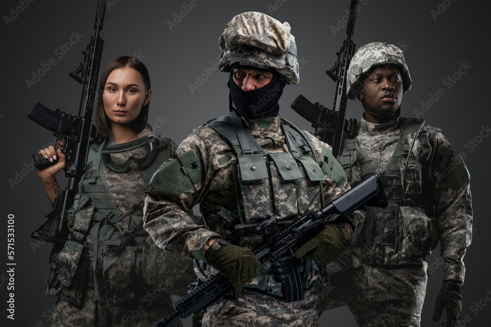 Multiethnic military team of three soldiers with rifles against grey background.