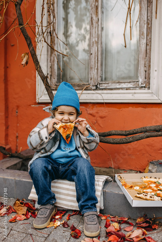 Little happy boy eating pizza, outdoors, sitting on city street.