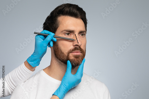 Doctor in protective gloves drawing marks on man's nose for cosmetic surgery operation, grey background