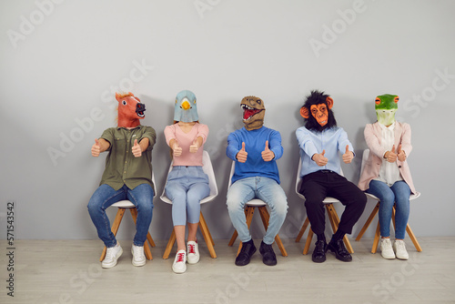 Group of young people in trendy rubber masks of different animals' heads showing thumbs up. People in masks of horse, pigeon, dinosaur, monkey and frog sit in row on chairs against gray wall.