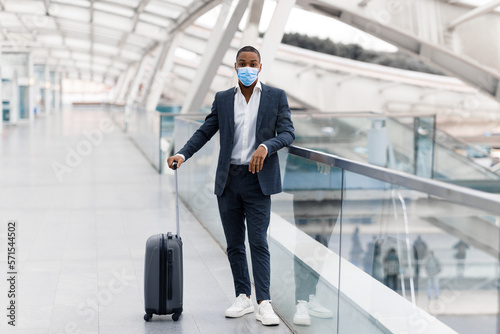 Black Man Wearing Suit And Medical Mask Posing With Suitcase At Airport