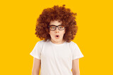 Portrait of emotional teen boy wearing curly brown wig. Amazed teenager looking at camera rounding lips shocked with unexpected news. Frightened boy standing on isolated yellow background