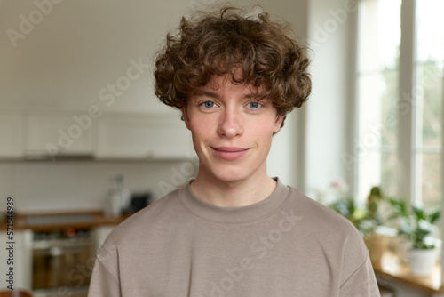 Close up of a young handsome positive curly man with blue eyes with a smiling face wearing casual beige t-shirt looking at camera and standing alone in morning in the modern kitchen at home.