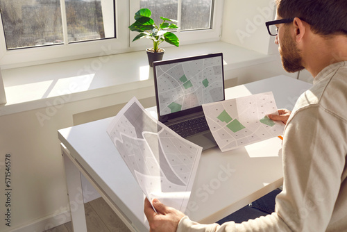 Serious engineer with cadastral maps and projects is sitting at desk with laptop in office. Cadastral engineer carries out accounting of land plots. Ownership of real estate object. photo