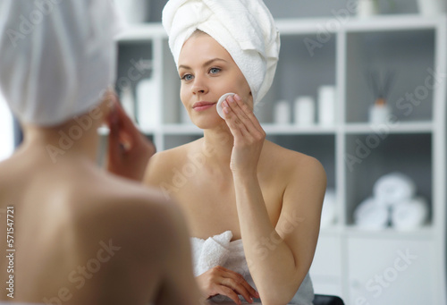 Young woman sits in the bathroom in front of the makeup mirror and does cosmetic procedures. Beautiful girl in white towel. Skin care  health  rejuvenation and spa treatment concept.