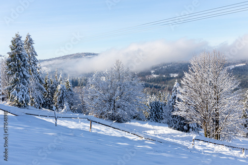 Winter landscape of Beskid Mountains in Poland, forest and mountain glade with wooden fence covered with fresh white snow, mountains and clouds in the background, Wegierska Gorka.