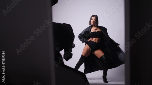 sexy young fashionable woman model in a black swimsuit and leather boots and coat is photographed in the studio with a photographer on a white background. backstage