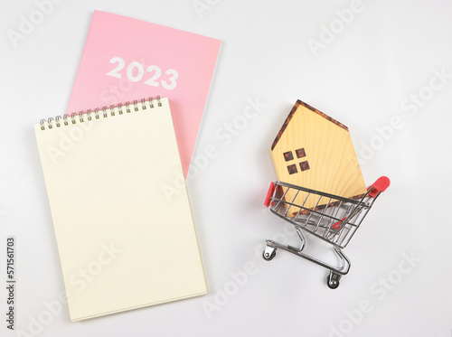 flat layout of wooden house model in shopping trolley with blank page notebook and pink diary or 2023 on white background with copy space, home purchase plan concept.