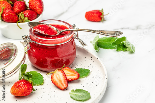 Strawberry jam in the glass jar with a spoon at white table. top view