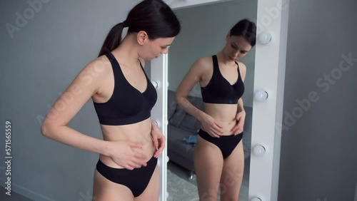 Woman in black underwear looking on her fat on her flabby belly in mirror, close-up of her waist. Skin care and weight loss concept. A young thin woman critically examines her figure in the mirror. photo