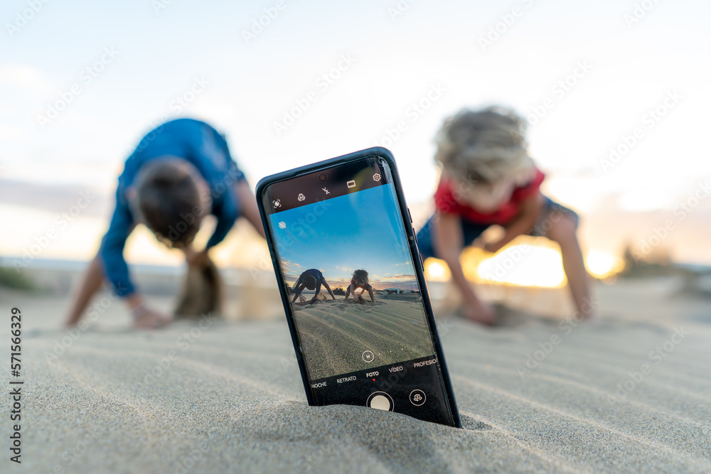 View of children playing on a sandy beach through a smartphone screen during sunset. - obrazy, fototapety, plakaty 