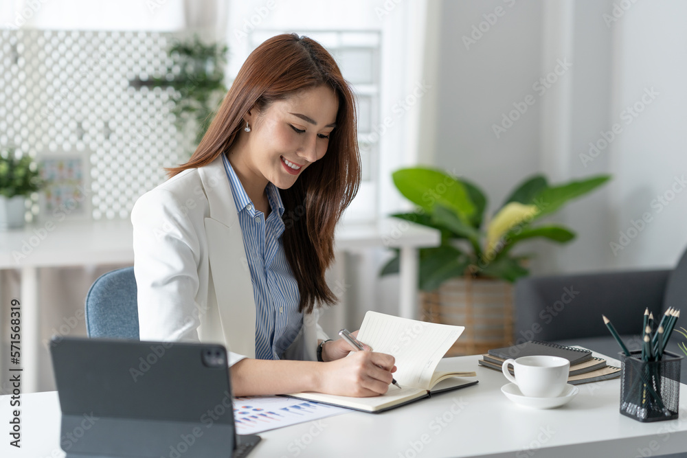 Confident Asian businesswoman sitting and taking notes in financial book Income tax with laptop computer or tablet in a happy office. Confident Asian businesswoman sitting and taking notes in financial book Income tax with laptop computer or tablet in a happy office.